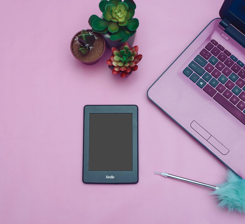 Flat lay of a modern desk setup with Kindle, laptop, and succulent plants on a pink background.