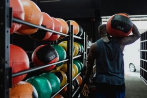 Back view of a man carrying a 20LB medicine ball in a gym with colorful balls on racks.