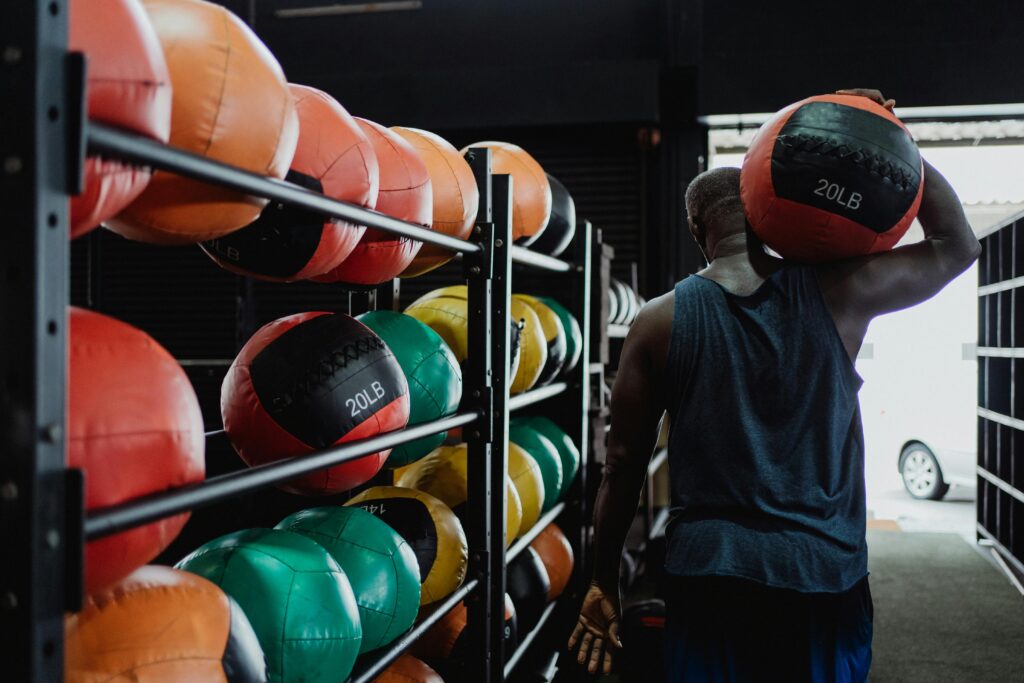 Back view of a man carrying a 20LB medicine ball in a gym with colorful balls on racks.