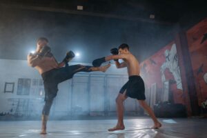 Two male fighters engage in an intense martial arts sparring session indoors with dramatic lighting.
