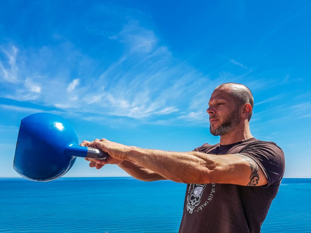 A fit, tattooed man lifting a kettlebell outdoors by the ocean under a clear blue sky.