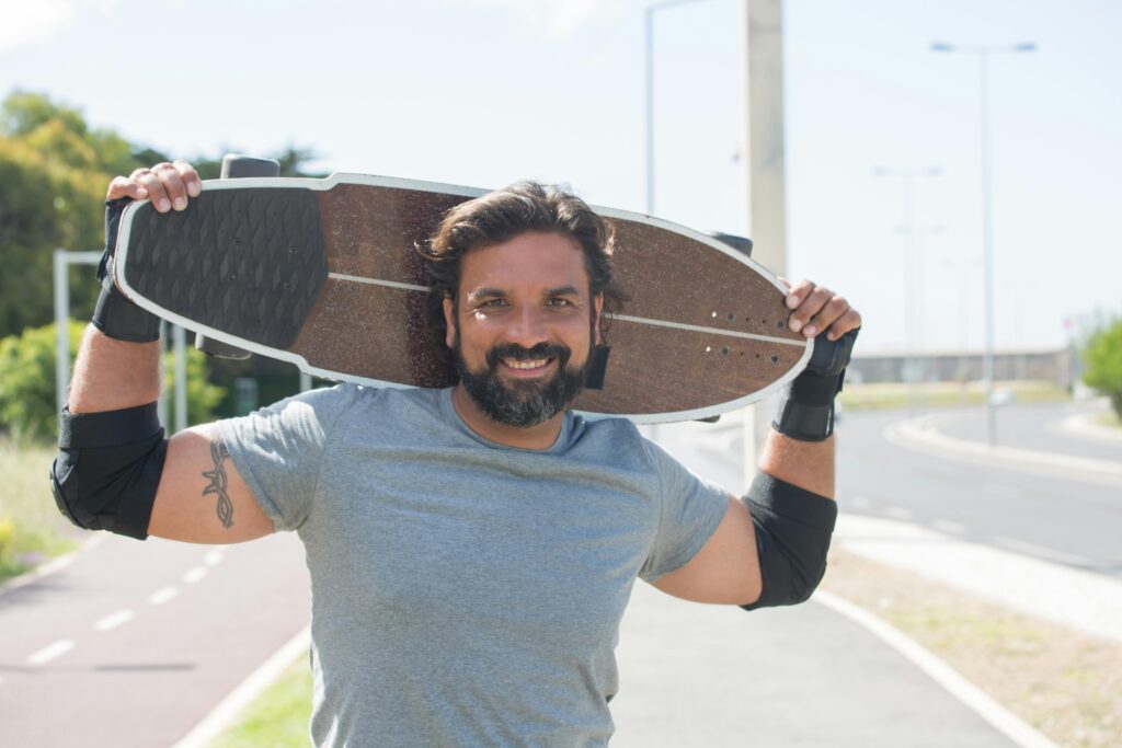 Smiling man holding a longboard on a sunny day, enjoying outdoor activities.