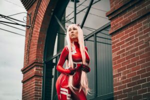 A cosplayer in a red costume poses confidently against a brick building.