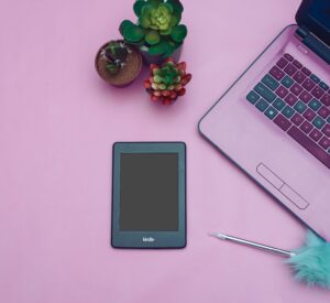 Flat lay of a modern desk setup with Kindle, laptop, and succulent plants on a pink background.
