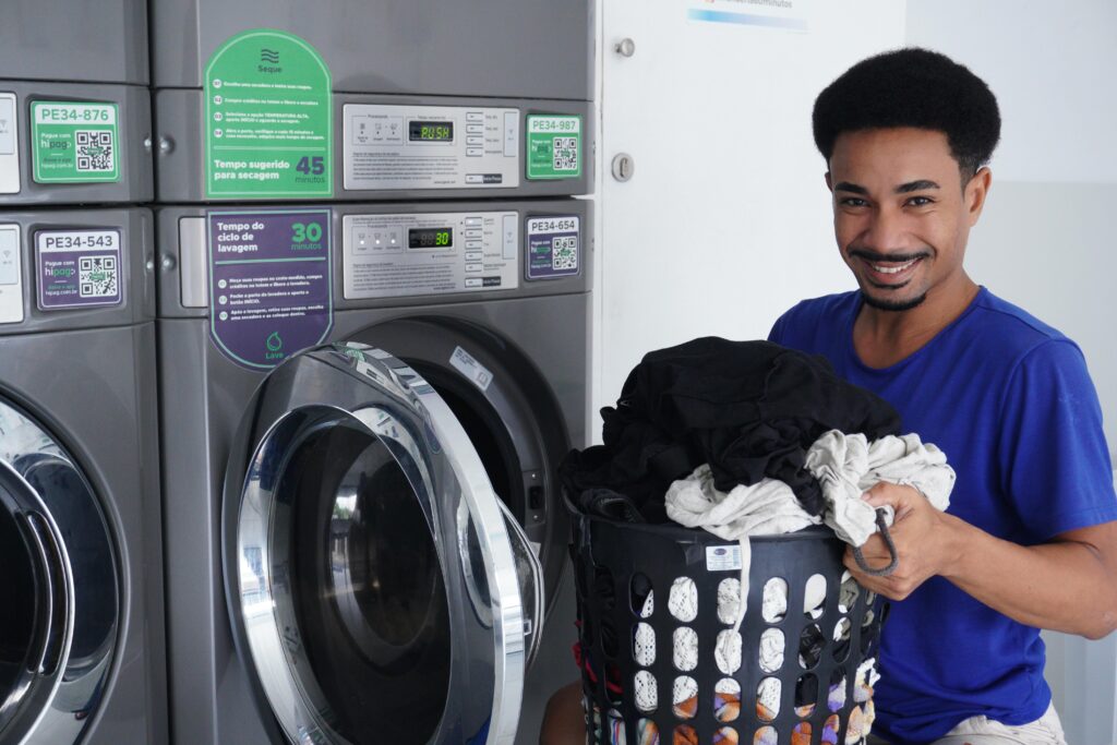 Smiling man holding laundry basket in a laundromat, ready to wash clothes.