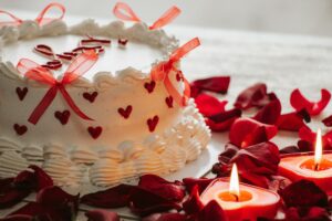 Heart-decorated cake with red bows, candles, and rose petals for a romantic setting.