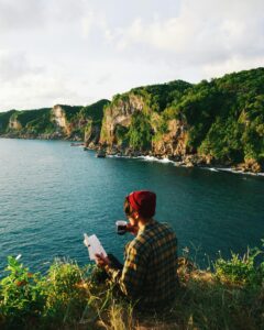 Person enjoys a peaceful moment, reading by the ocean on a cliffside, surrounded by nature.