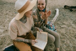 Two women with tattoos enjoying a book on a sandy beach, sharing smiles and clapping.