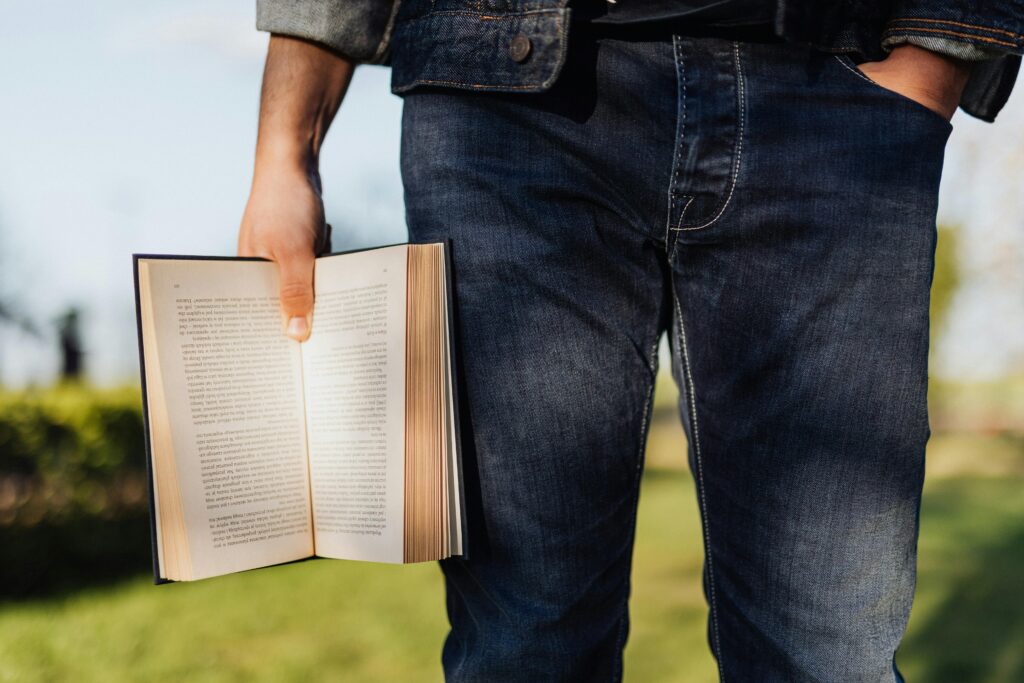 Young man casually holding an open book outdoors in a sunny, peaceful park setting.