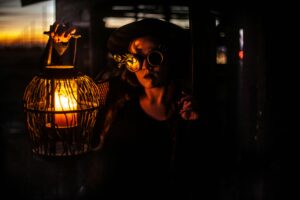 A woman with steampunk goggles holds a lantern, creating an eerie glow in a dark setting.