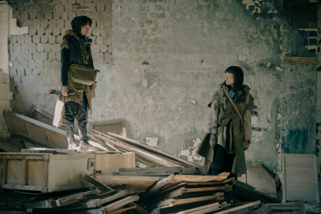 Two people exploring a desolate, abandoned building in post-apocalyptic attire.