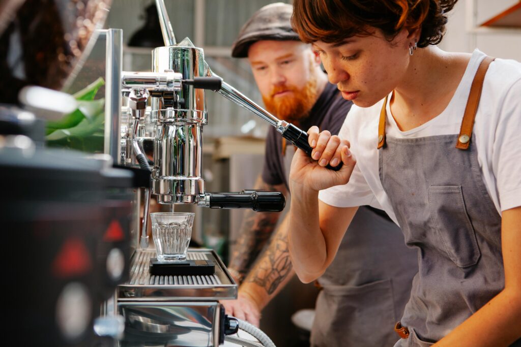 Crop female employee touching lever of professional coffee maker pouring water into glass against bearded partner in cafeteria