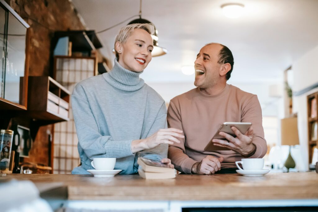 Delighted adult ethnic man laughing and looking at wife while watching together funny video on tablet and drinking coffee in sunny morning at home