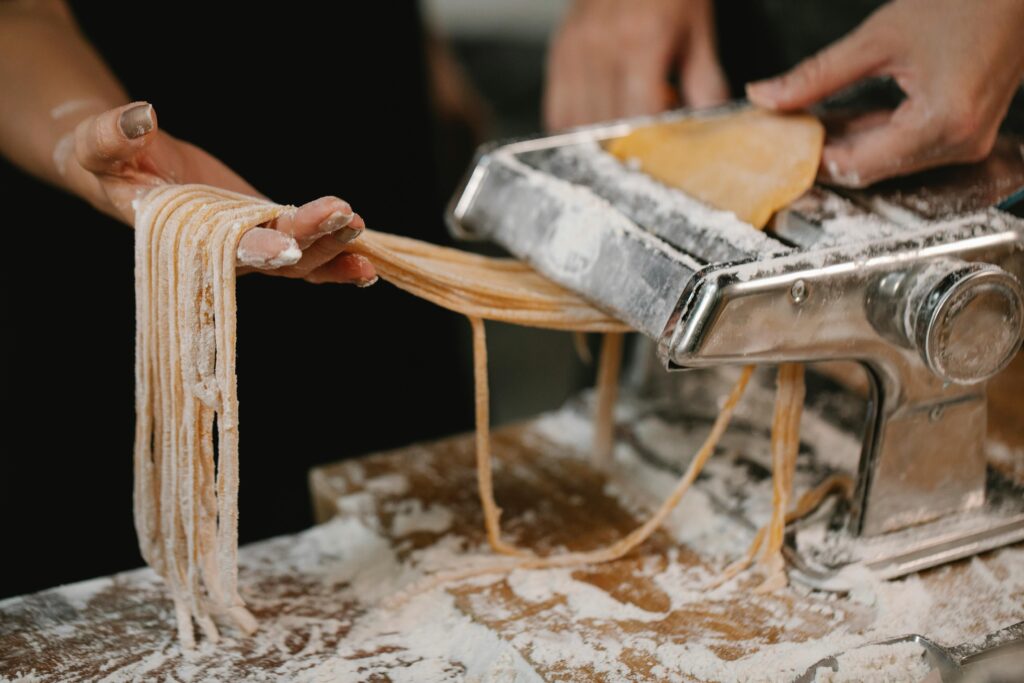 Crop anonymous female producing long noodle with metal pasta cutter on wooden table covered with flour
