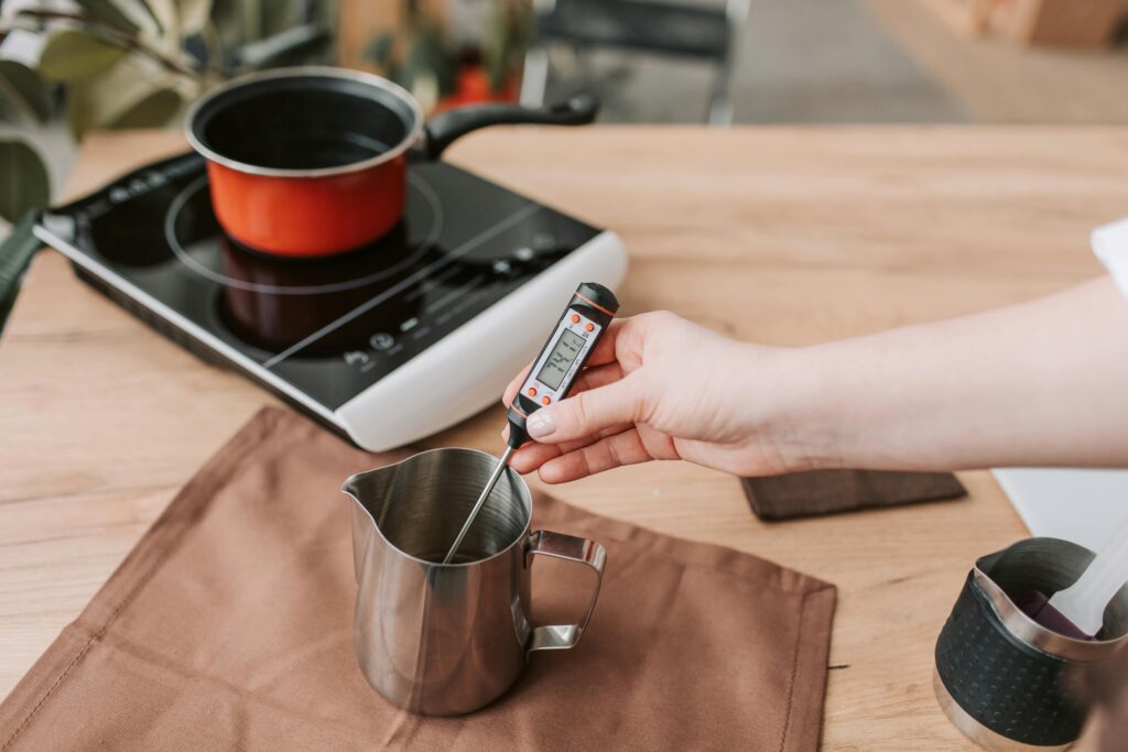 A person measures the temperature of melted wax on an electric stove, highlighting modern craft techniques.