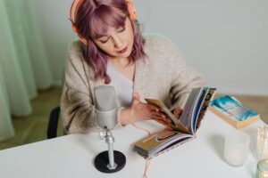 Young woman podcasting at a desk with headphones and books, using a microphone.