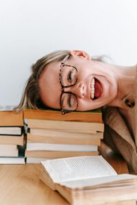 Smiling woman with glasses resting her head on a stack of books, exuding happiness.