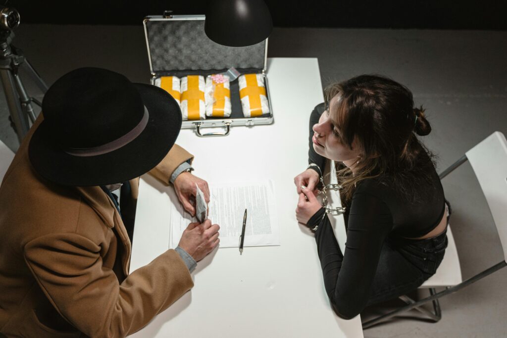 Two individuals engage in a tense negotiation with a briefcase of suspicious packages in a dimly lit room.