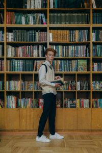 A young man in a sweater vest browsing books in a library.