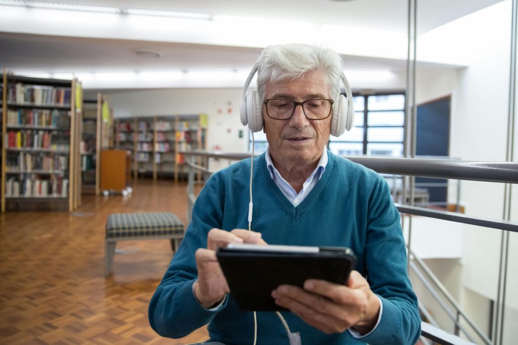 Senior man wearing headphones, using a tablet in a library setting, engaged in digital learning.