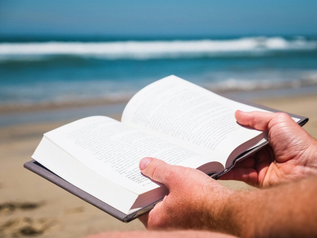 Hands holding an open book at a beach with waves in the background, perfect for summer relaxation.