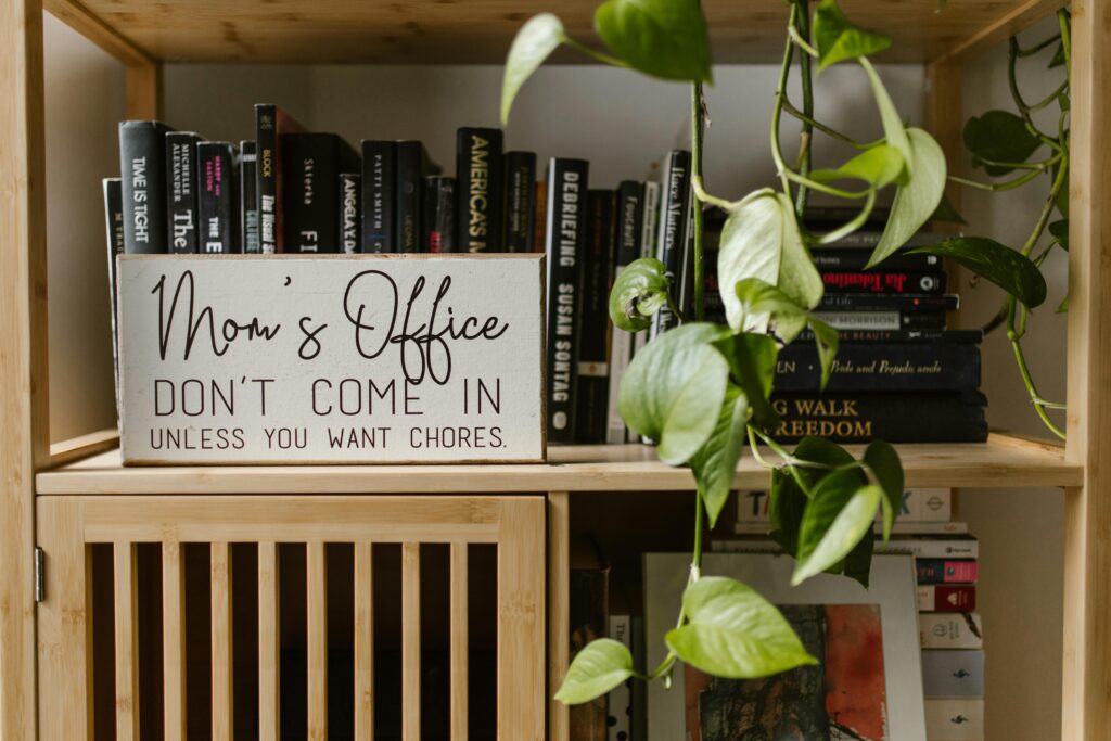 A wooden bookshelf filled with books and plants, with a humorous sign in a home office setting.