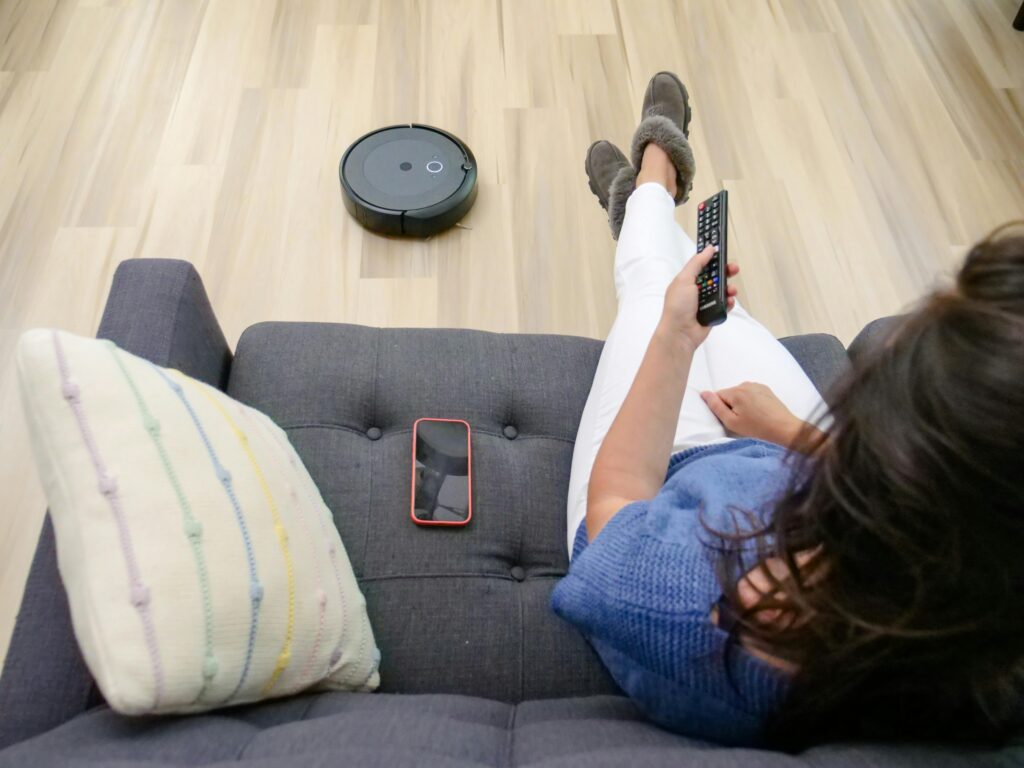 Person relaxing on a sofa controlling a robot vacuum cleaner with a remote.