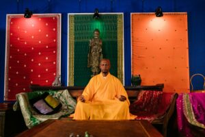 A Buddhist monk in orange robes meditates indoors, surrounded by colorful drapes and spiritual decor.
