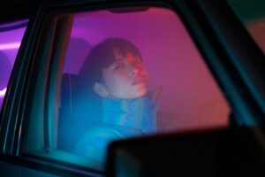A woman in a colorful jacket sits inside a car at night, illuminated by neon lights.