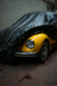 A vibrant yellow vintage car partially draped with a black cover in an urban alleyway.