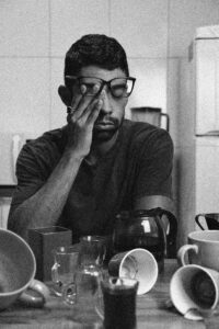 A tired man wearing glasses sits at a table, surrounded by empty coffee cups.