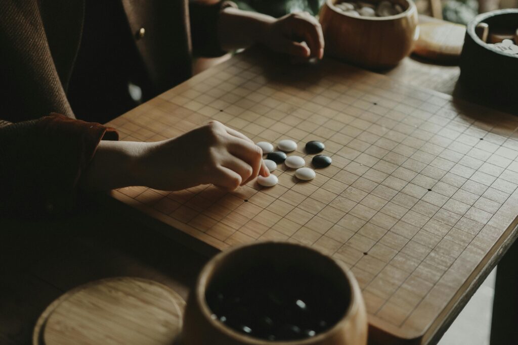 A person playing Go, arranging black and white stones on a wooden board indoors.