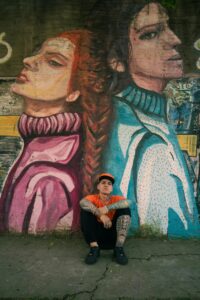 Young man in orange cap sits by colorful street mural with artistic portraits.