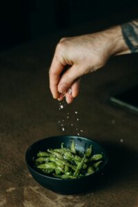 A hand sprinkles salt over a bowl of fresh edamame beans, capturing a moment of delicious meal preparation.