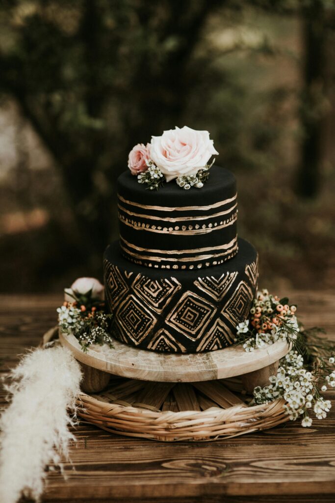 A stunning black and gold wedding cake with floral decorations on a wooden table outdoors.