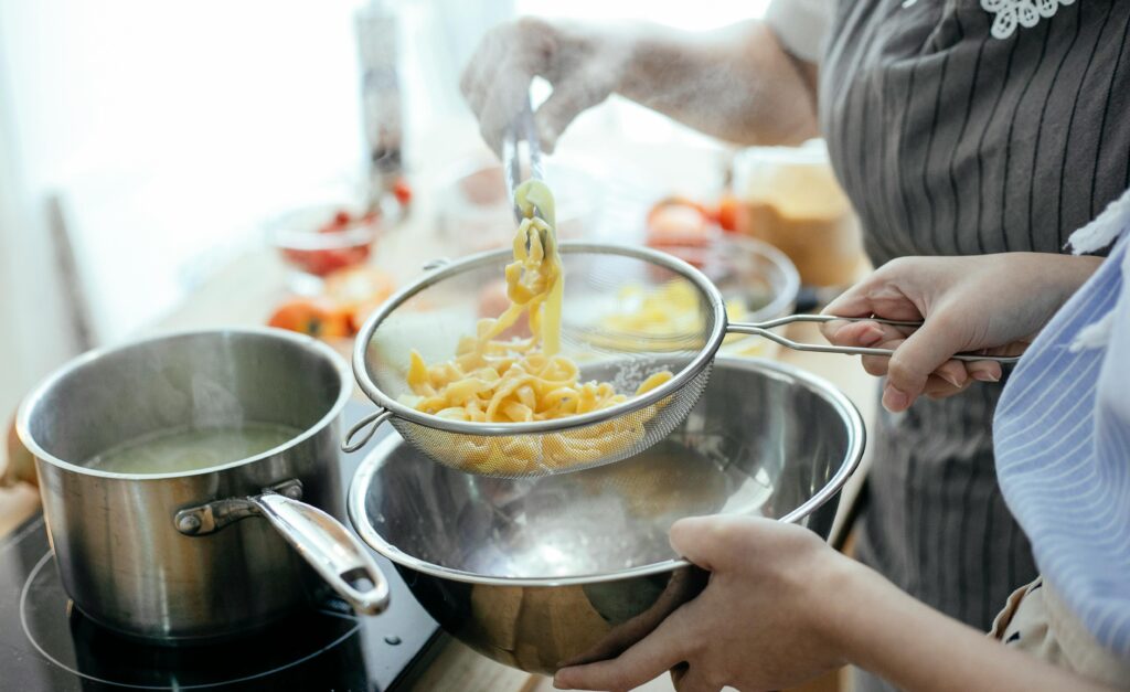 High angle of crop faceless female chefs putting noodles from saucepan with boiling water into strainer while standing at table with ingredients in kitchen