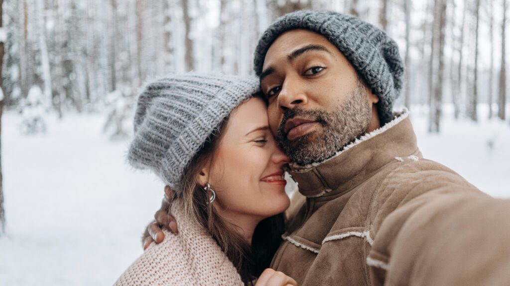 A joyful couple embracing while taking a selfie in a snowy winter forest, showcasing love and diversity.