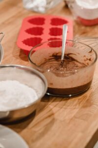 Glass bowl with chocolate batter placed on wooden table with silicone molds and flour in kitchen at home during cooking process