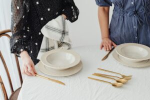 Two women prepare an elegant table setting with gold cutlery and bowls on a white tablecloth.