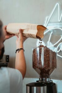 Barista pouring coffee beans into grinder from a paper bag inside a cafe.