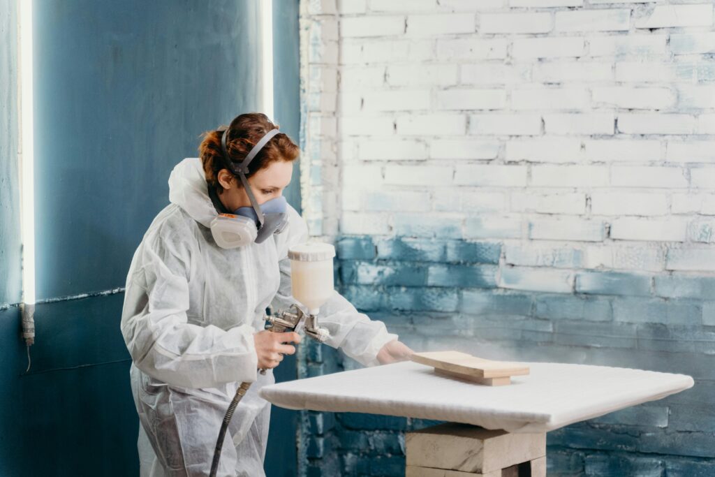 A female artisan wearing protective gear spray paints a wooden object indoors.