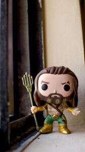 Close-up of an Aquaman Funko Pop figure by a window, highlighting its detailed trident and pose.