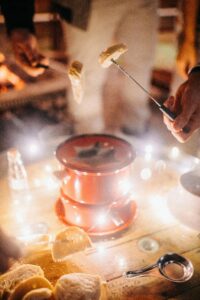 Crop anonymous friends melting cheese in fondue and eating bread while standing around wooden table with garland at night