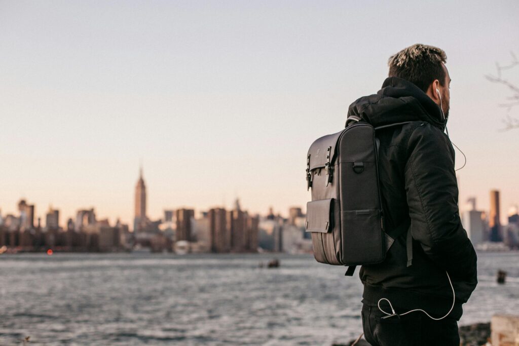 A man wearing a backpack gazes at the Manhattan skyline from Brooklyn waterfront during the day.