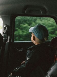 A young boy in a car gazes out a rain-streaked window, wearing a blue cap.