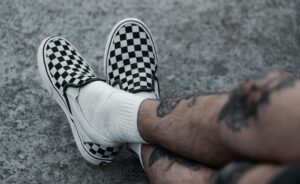 Close-up shot of tattooed legs wearing checkered slip-on shoes on textured ground.