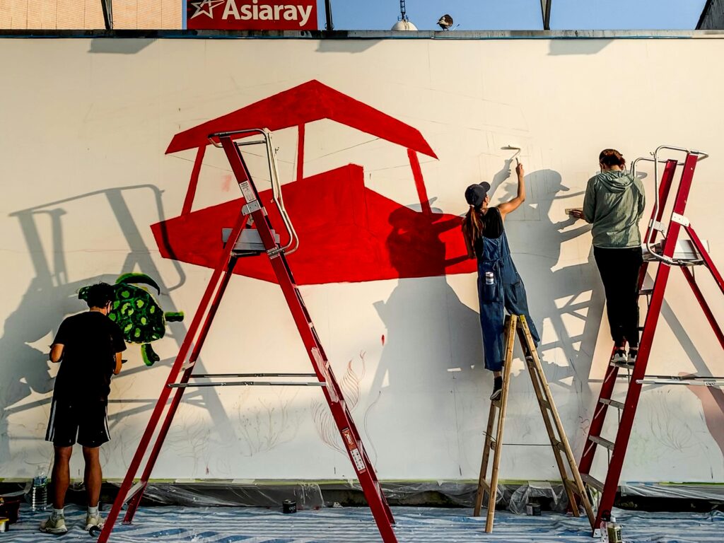 Three artists creating a colorful mural on a wall using ladders in an urban setting.