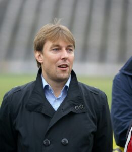 A man in a black coat smiles at an outdoor stadium, suggesting a sports event.