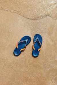Pair of blue flip flops on golden sand with gentle waves in the background.