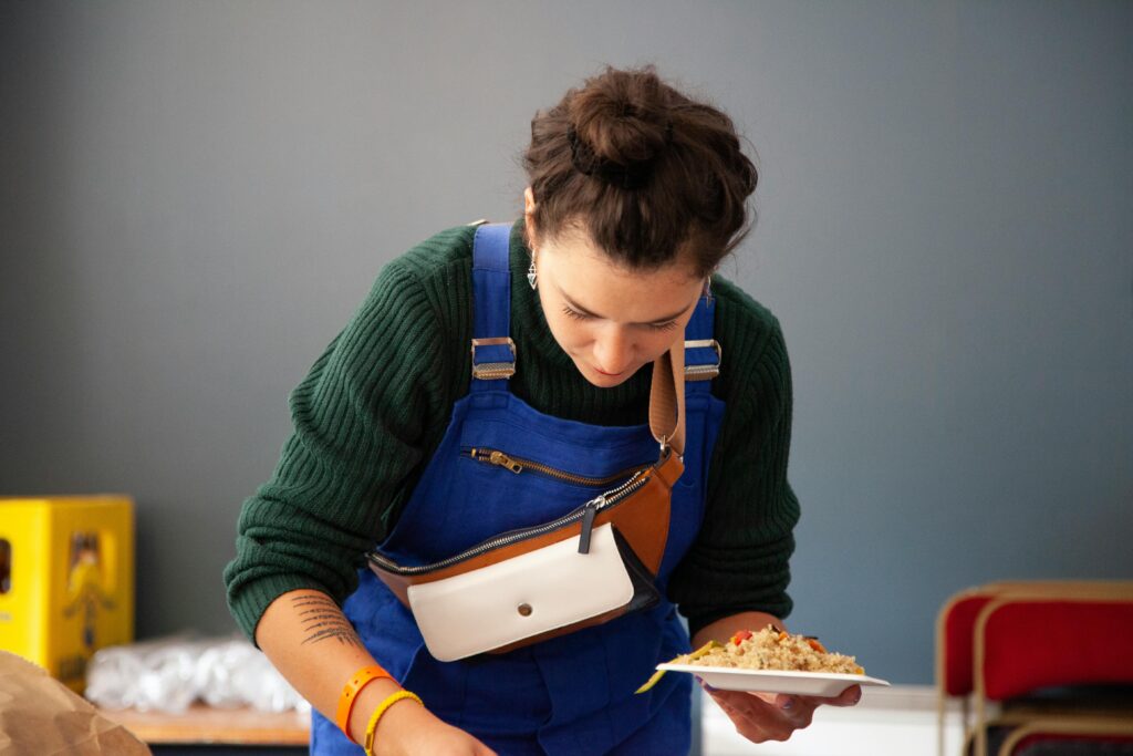 A young woman in blue overalls prepares food on a plate indoors, focused on cooking.